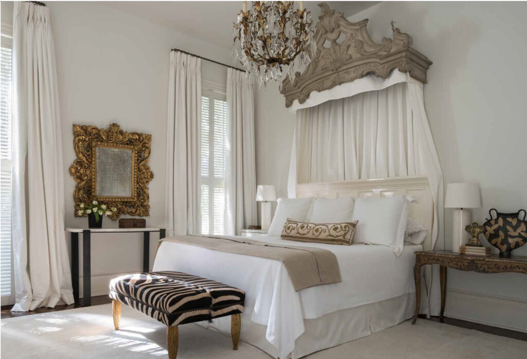 bedroom with white walls and white bed with antique french crown above and antiques in the room