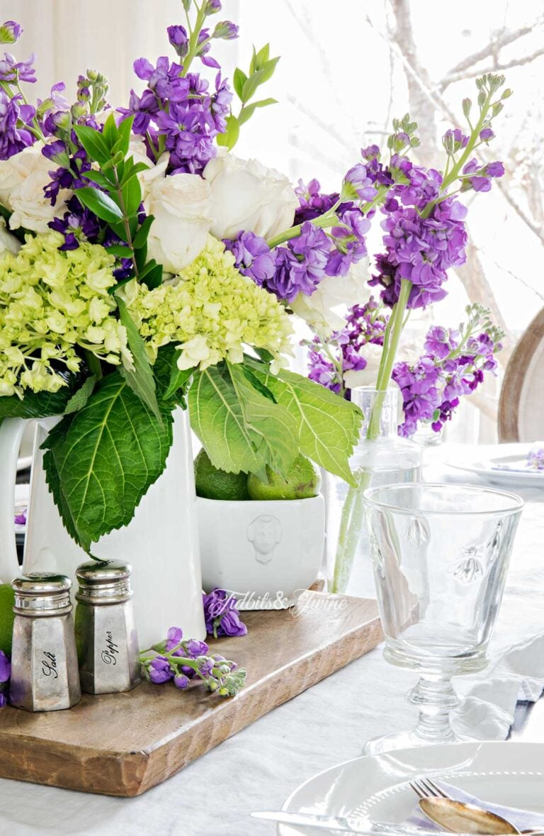 purple white and green flowers in a white pitcher on a dining table set with white dishes and purple gingham napkins