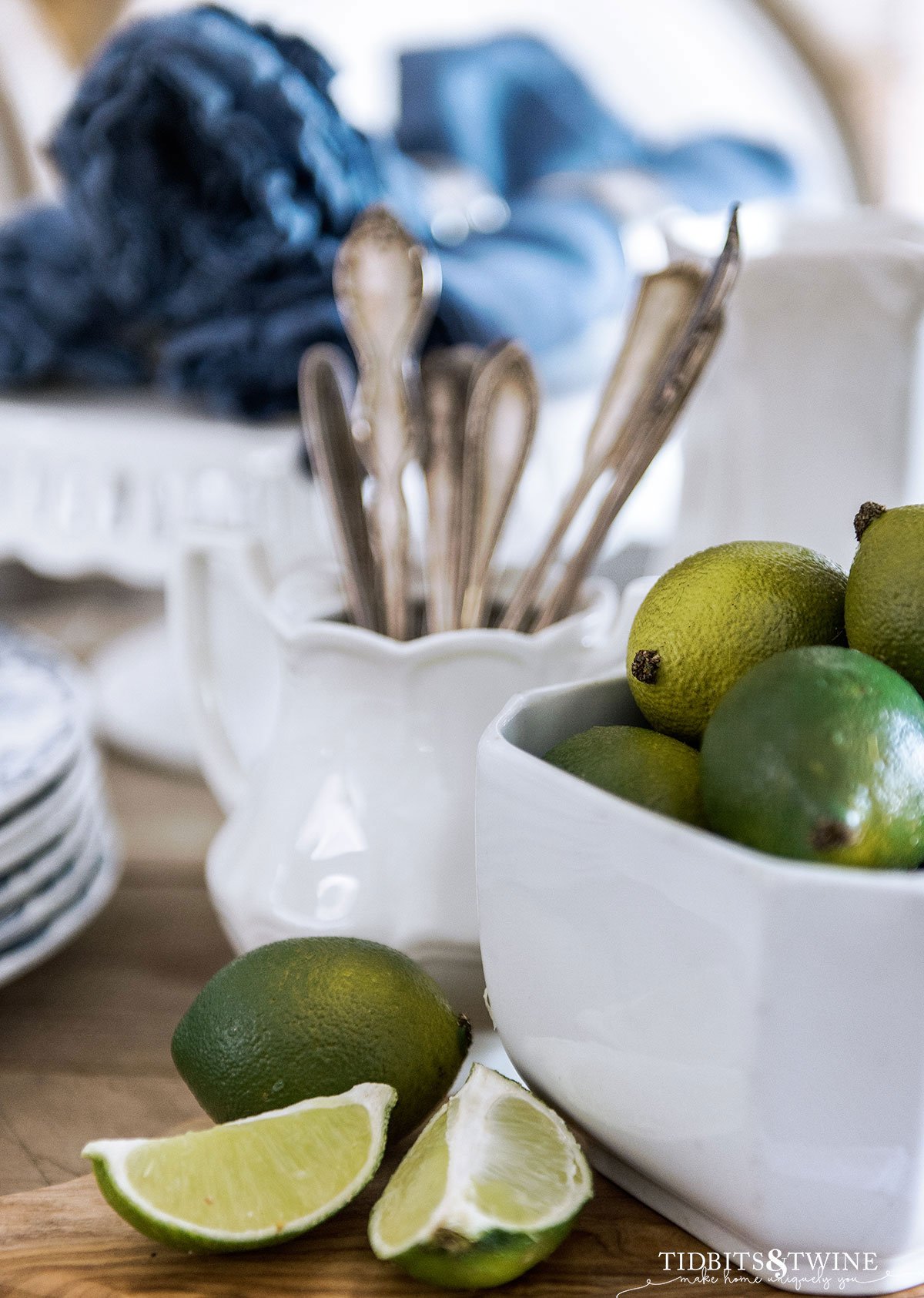 limes in a white ironstone pot next to one cut lime on cutting board and flatware in a sugarbowl