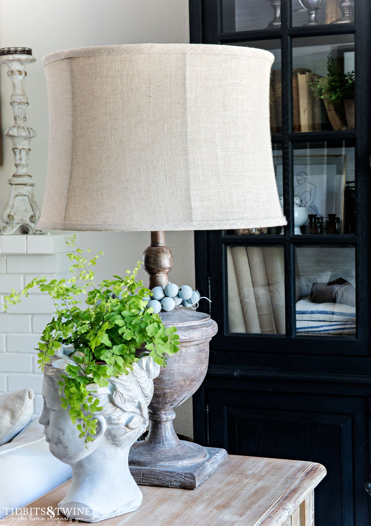 wooden lamp on sofa table with blue beads around base next to cement goddess head with a fern in it