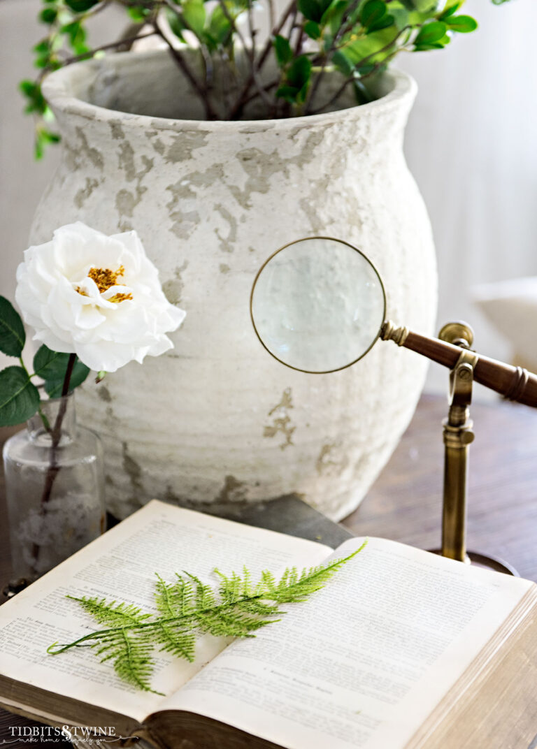 large rustic white vase with greenery behind open book with fern on top