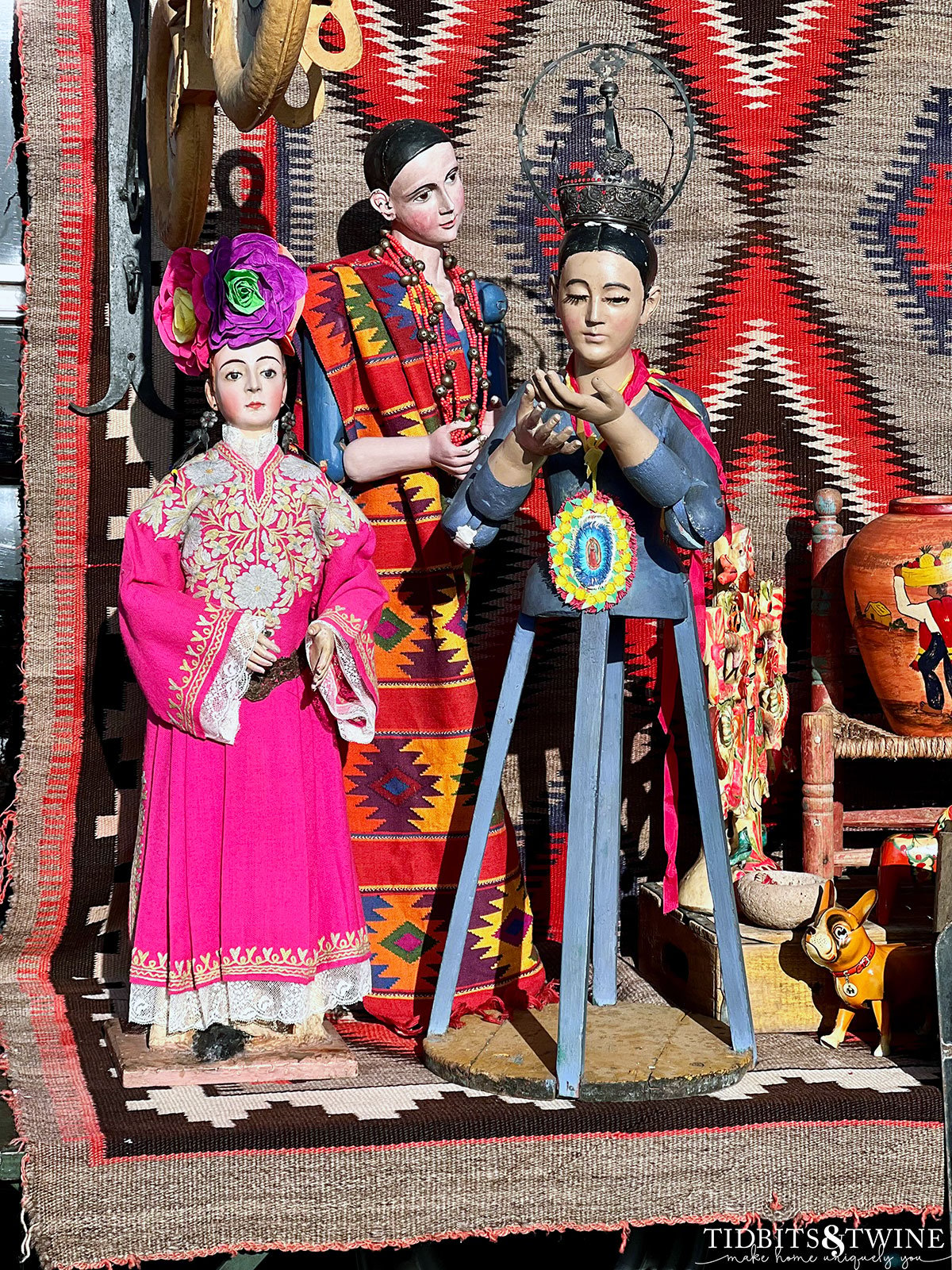 three religious santos dolls in blue and pink on table with kilm rug hanging in background