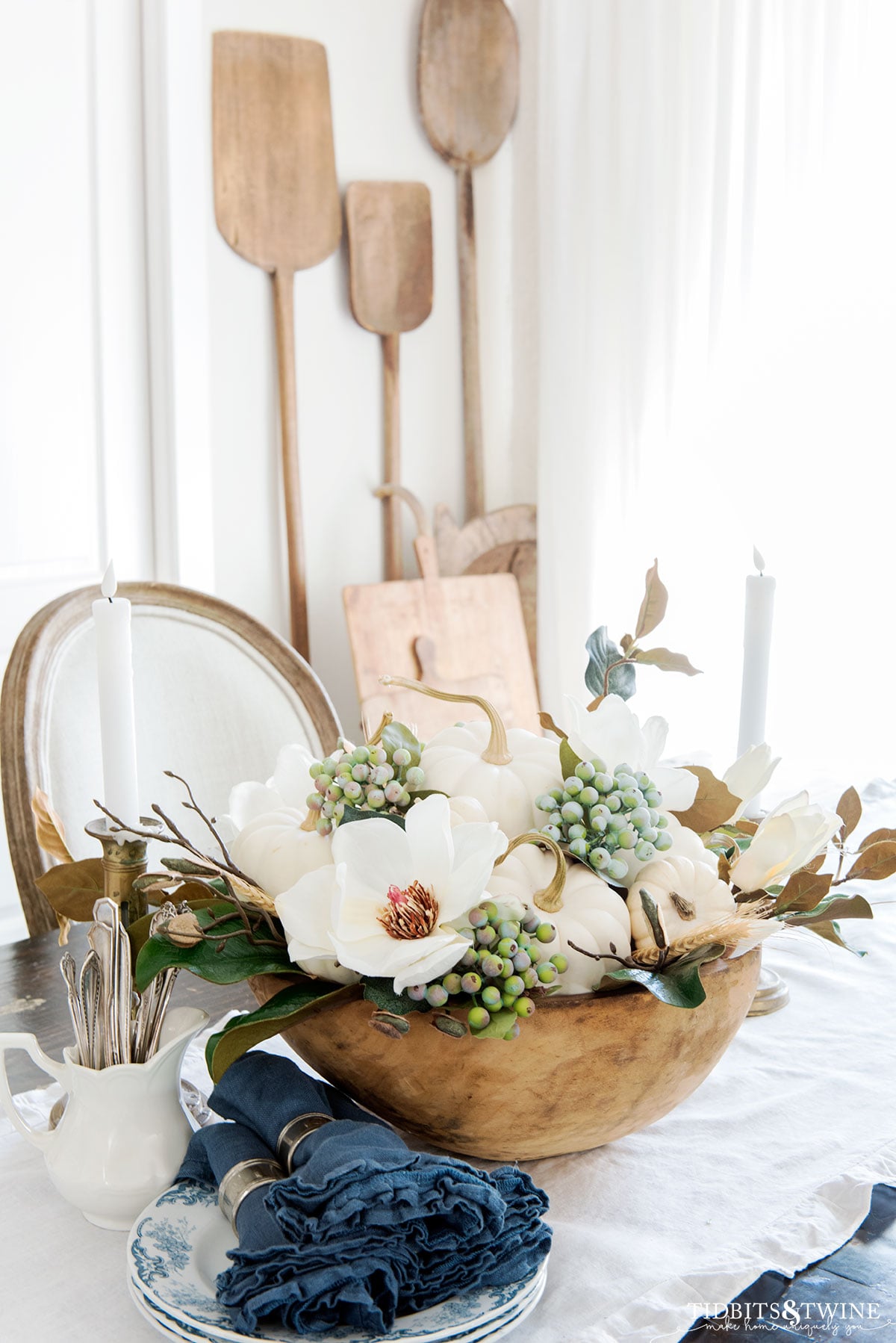 Antique wooden dough bowl filled with white pumpkins, magnolia flowers, and berries as a fall dining table centerpiece with blue linen napkins.
Copy alt text