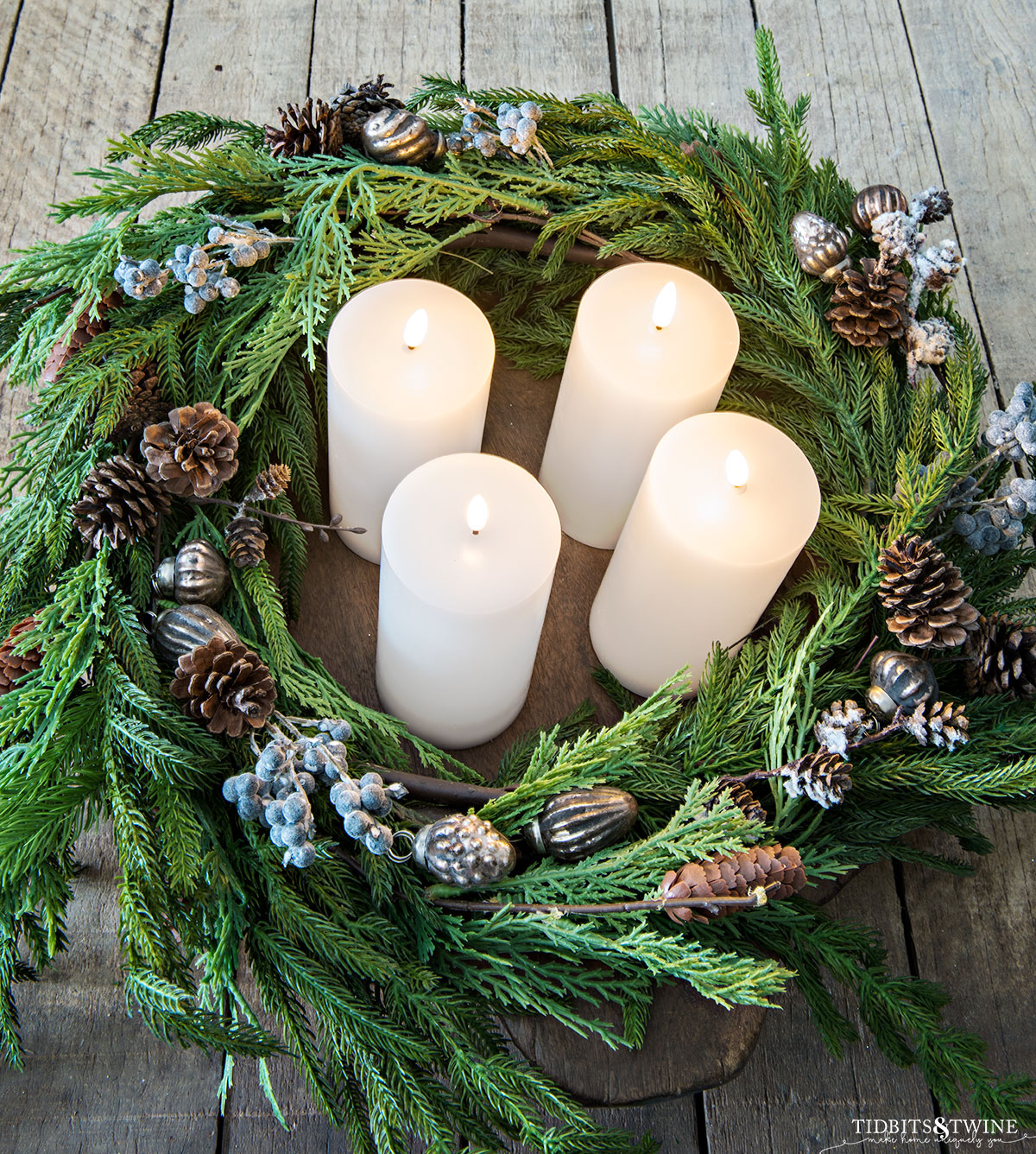 round dough bowl with norfolk pine and cedar branches wrapped inside and four white pillar led candles in the middle with berries and pinecones