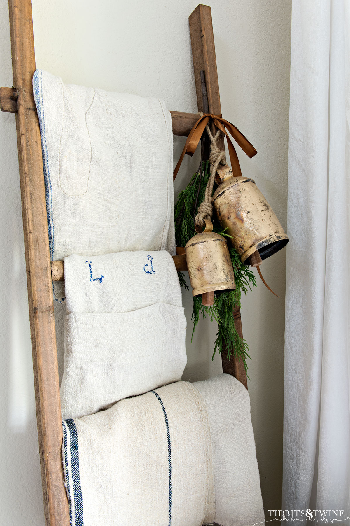 decorative wooden ladder in corner with antique grain sacks on rungs and rustic brass bells with greenery hanging from it