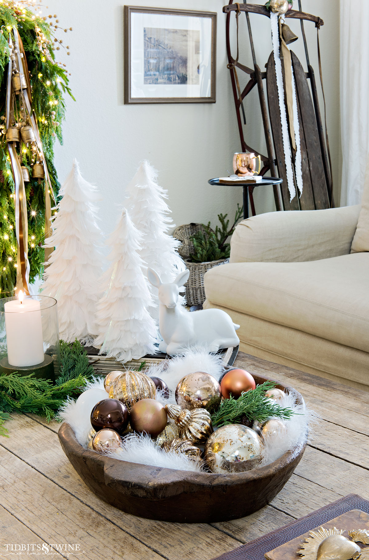 coffee table with round dough bowl full of gold and copper christmas ornaments with white feather trees in background