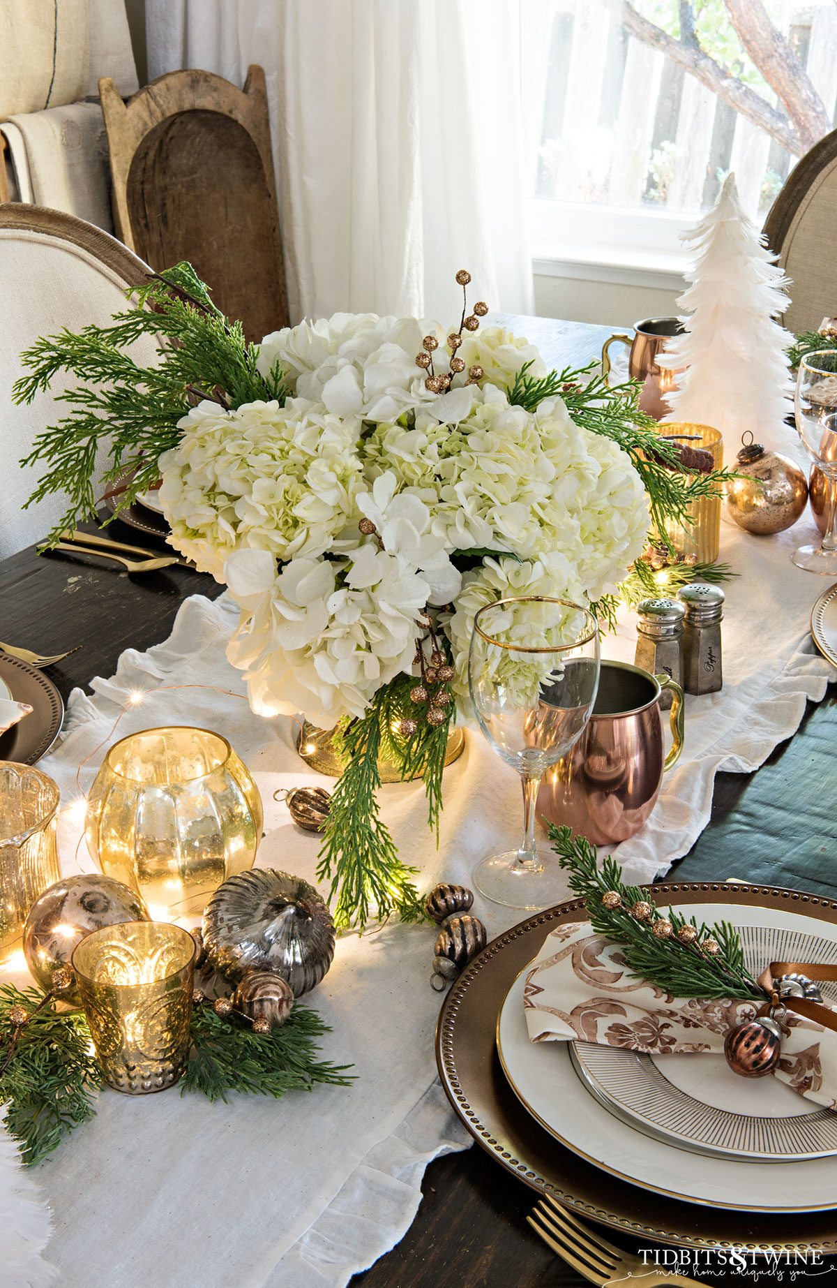 christmas tablescape with white hydrangeas and greenery in gold compote and votive candles and lights down center of table