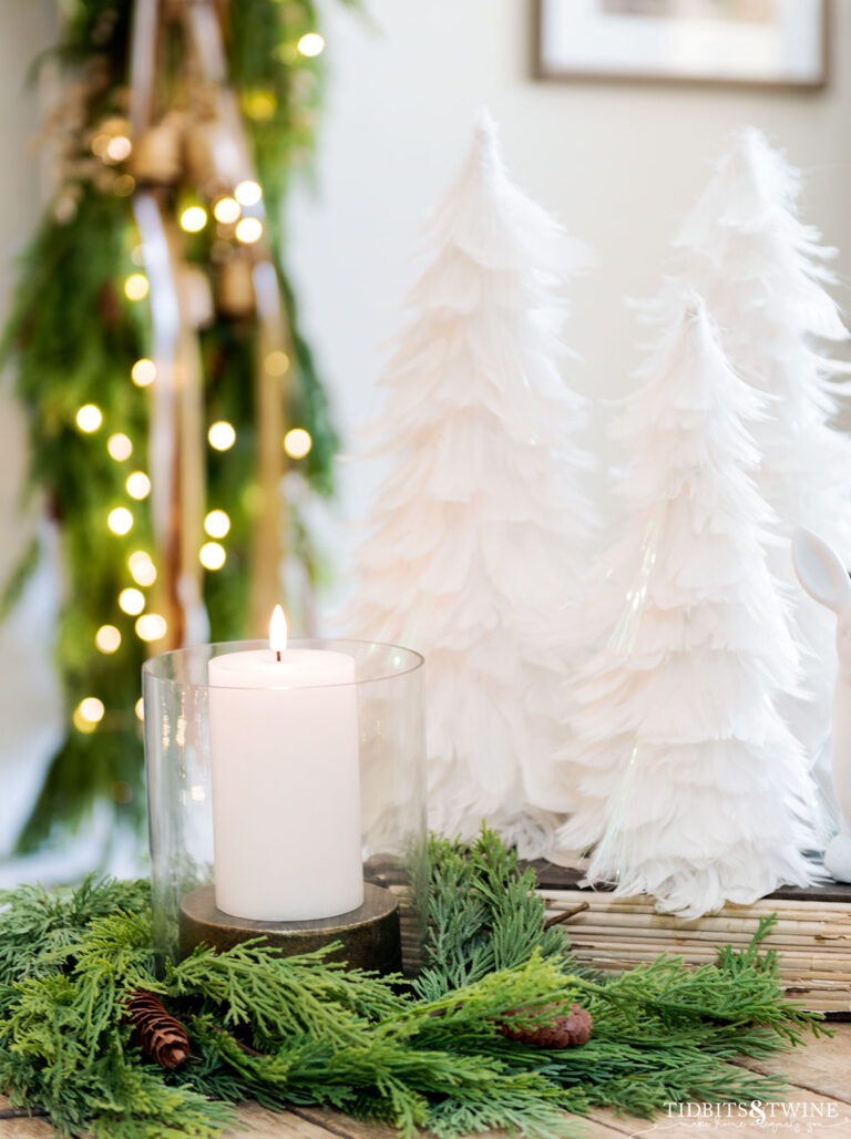 hurricane candle holder surrounded by christmas greenery with white feather christmas trees behind