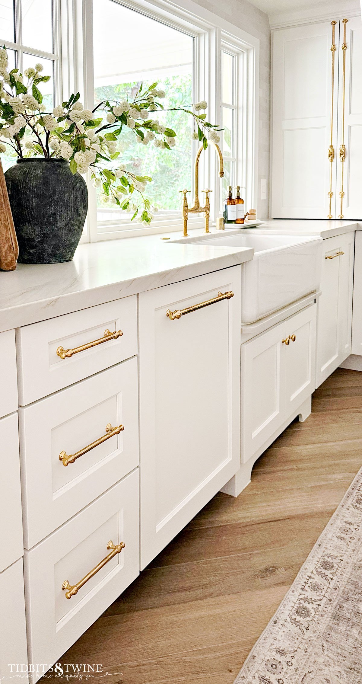 White kitchen cabinets with brass hardware and a farmhouse sink, styled with a large vase of greenery &mdash; showing how hardware and details complete a room.