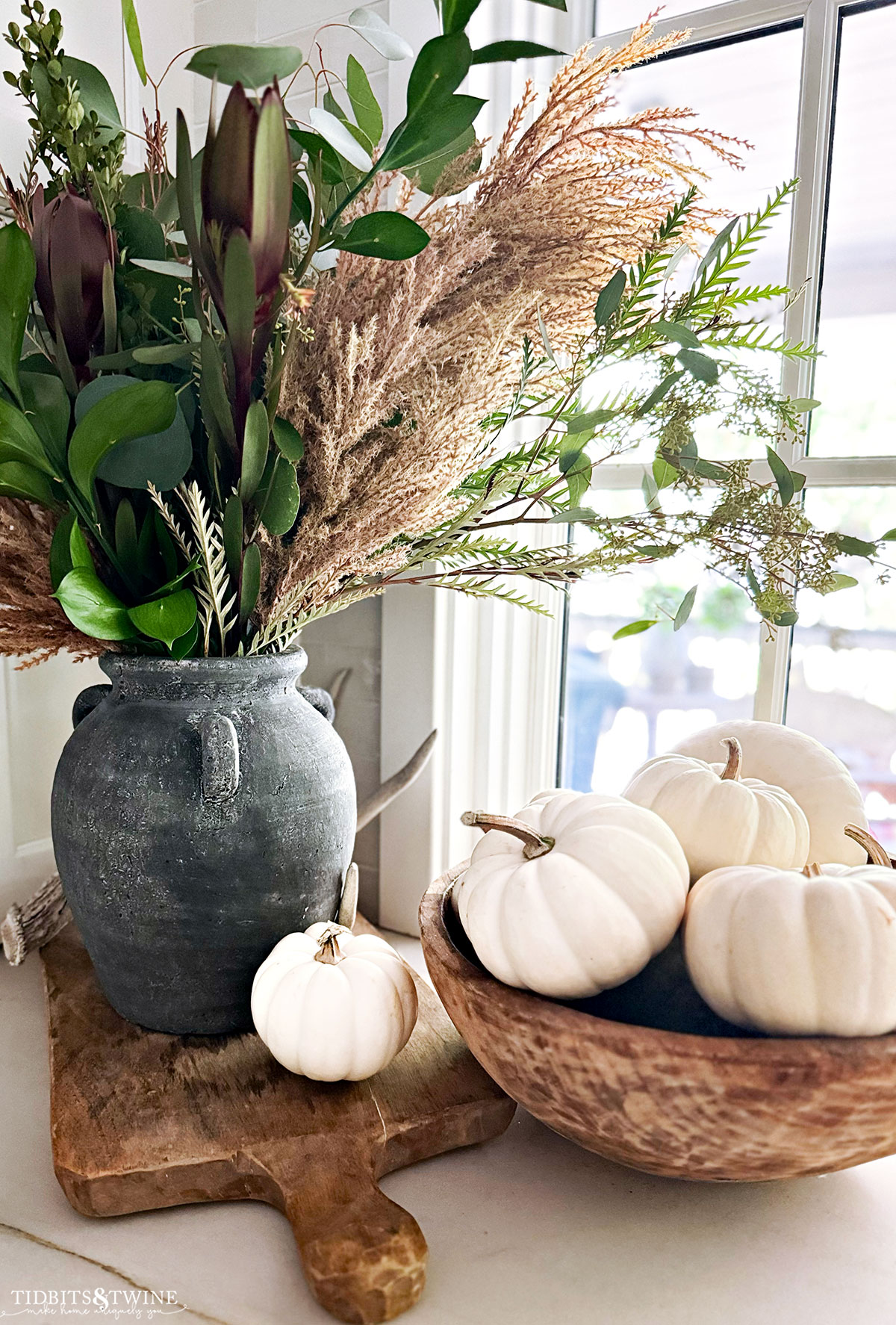 black vase in kitchen with greenery and pampas grass next to dough bowl of mini white pumpkins