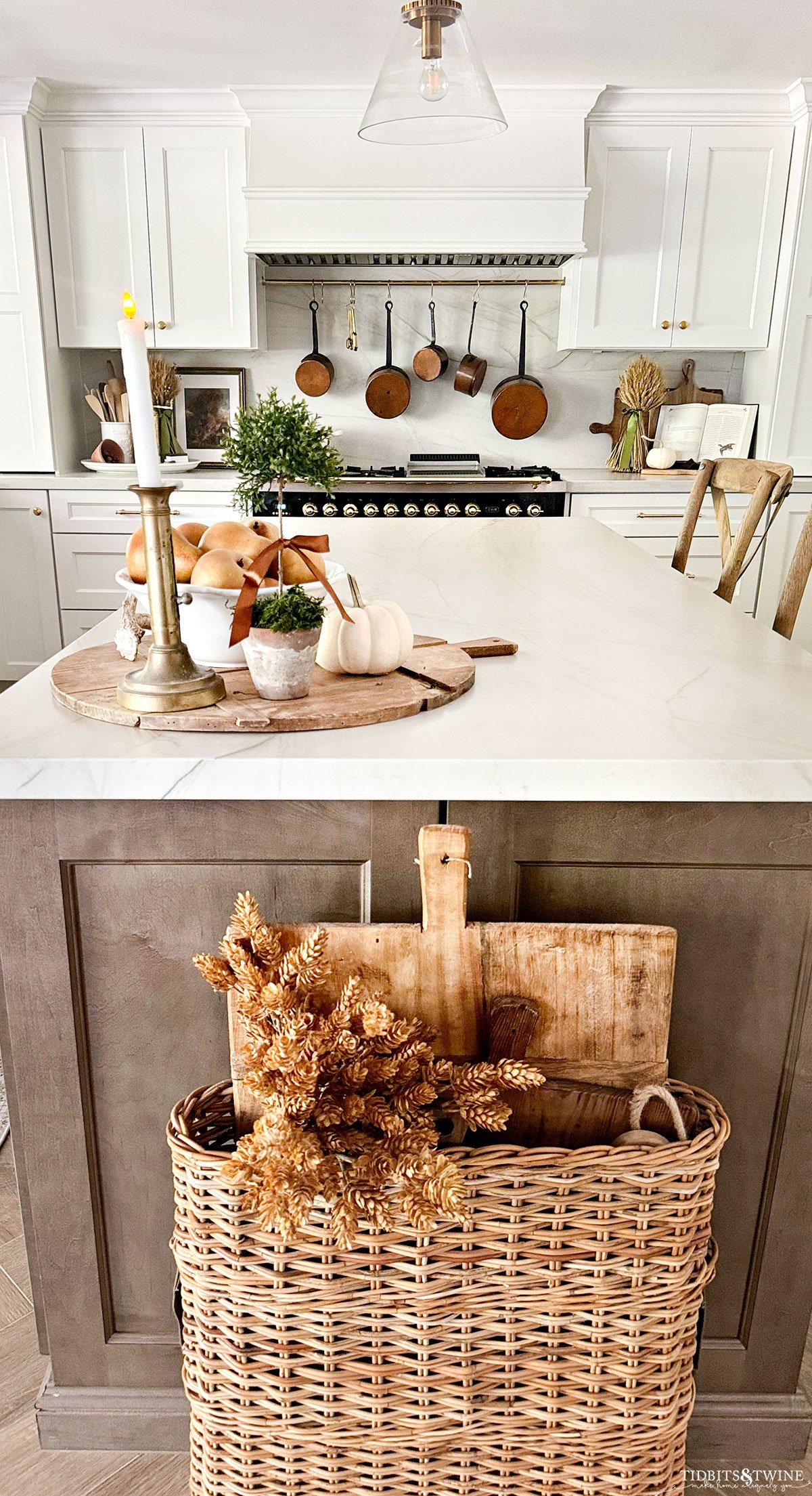 warm white kitchen decorated for fall with pears, pumpkins and wheat and large basket holding bread boards by island