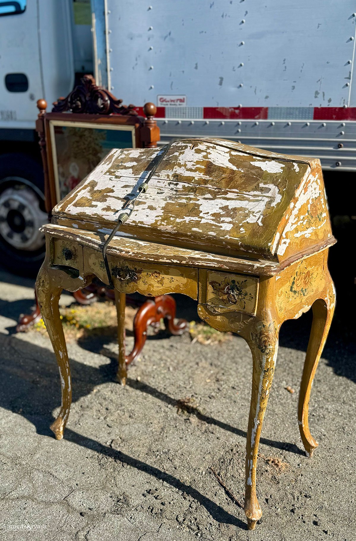 Chippy painted Italian desk with carved floral details, partially hidden beside a parked box truck.
