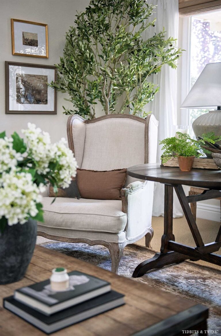 Living room chair beside an antique wine-tasting table with an olive tree and layered artwork