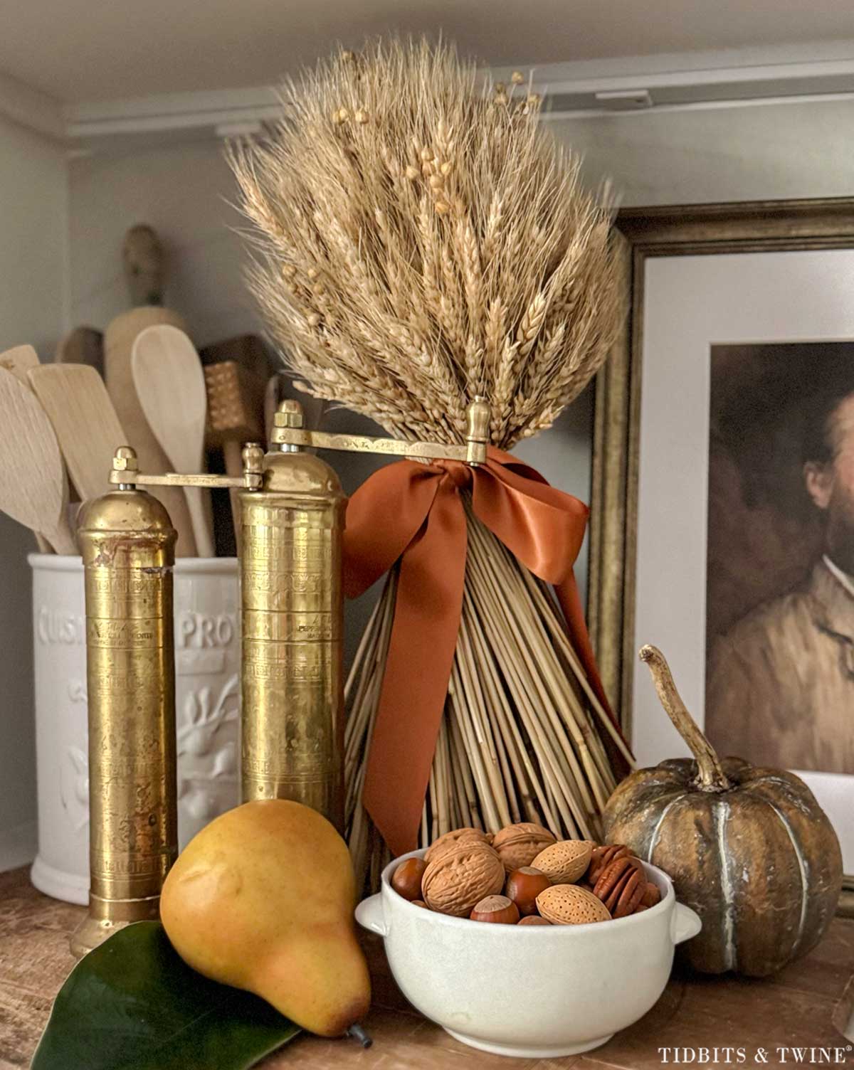 Walnuts and pecans in a small white bowl styled with pears, wheat, and brass kitchen accents