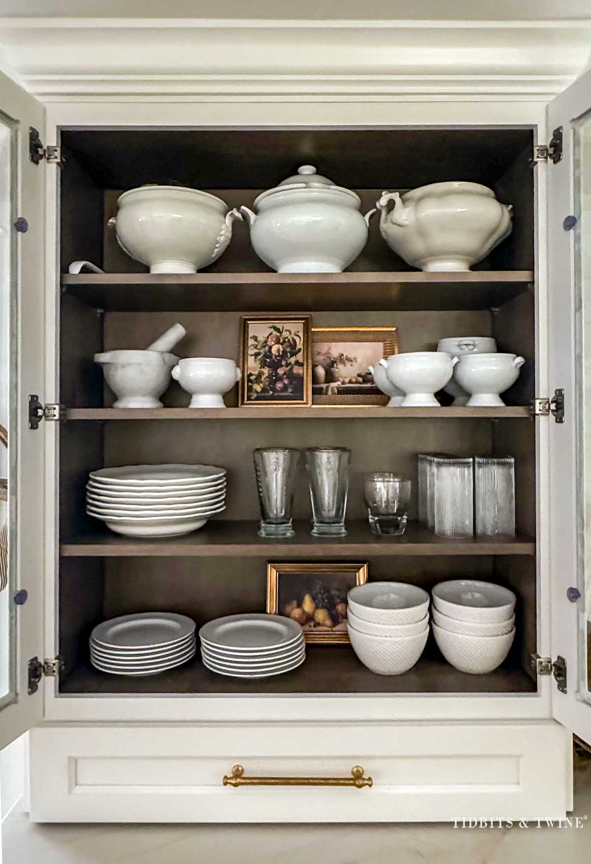 White dishes and small fall still life prints displayed inside a glass kitchen cabinet