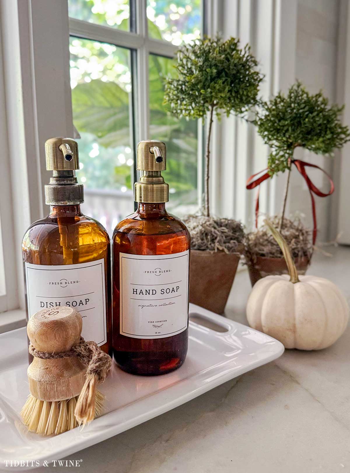 Amber soap bottles, wooden dish brush, topiaries and white pumpkin beside a kitchen sink