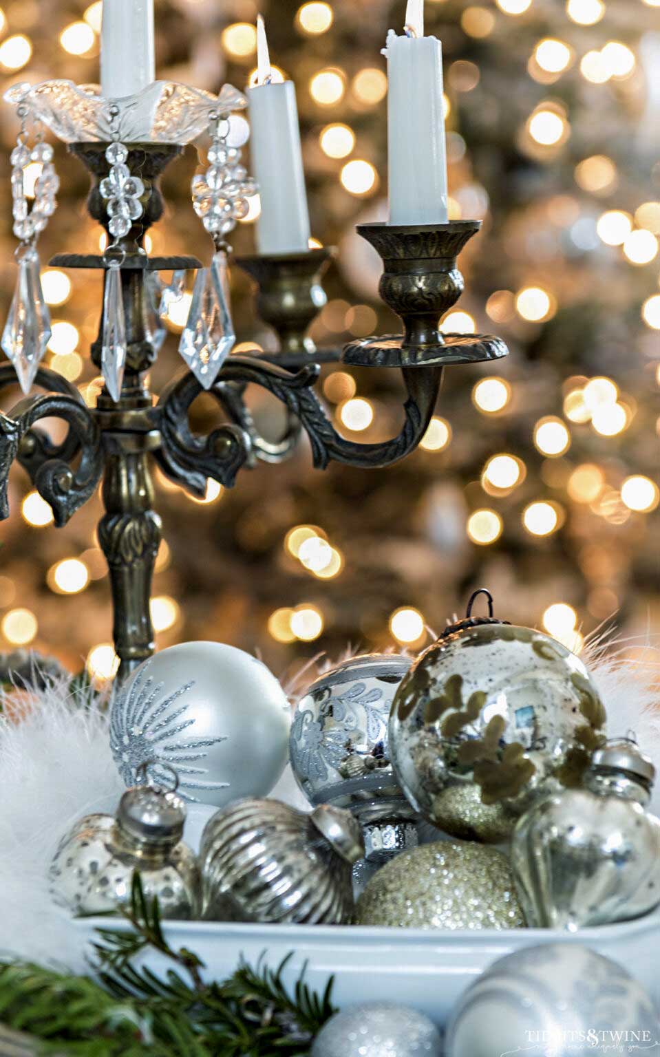 Vintage silver and mercury glass Christmas ornaments displayed beneath antique brass candelabra with softly glowing Christmas tree lights
Copy